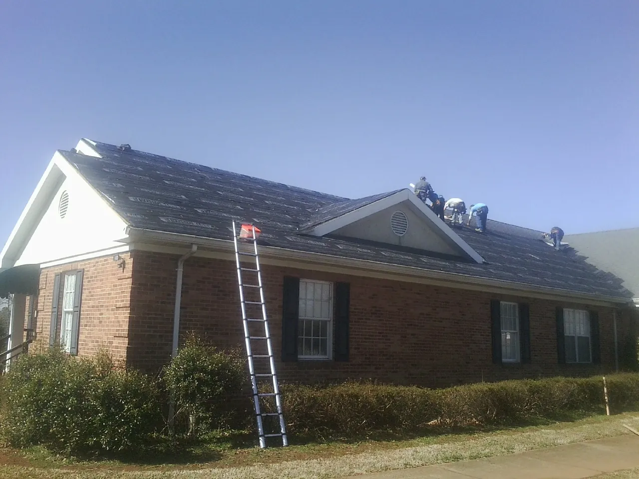 Crew tearing shingles off a brick ranch roof with a ladder set — Emergency Roof Repair work by our crew in Holly Ridge