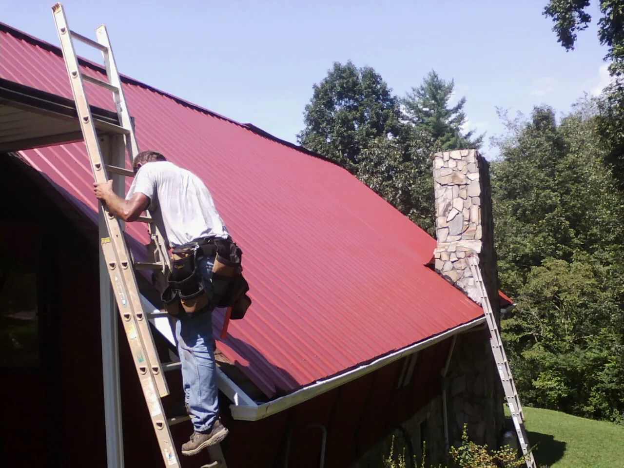 Roofer on a ladder beside a red metal roof and stone chimney — Soffit & Fascia Repair by Roofing Brothers in Holly Ridge